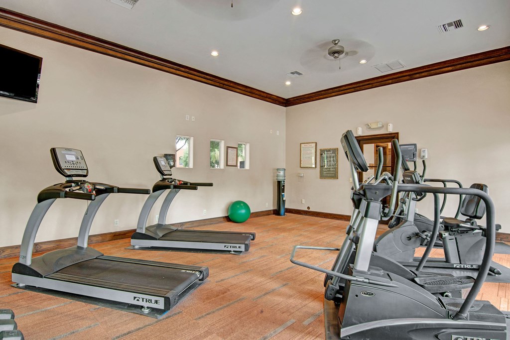 Modern gym interior here at The Retreat at Speedway Apartments with treadmills and elliptical machines on a wood floor. A green exercise ball and a television complement the calm, neutral tones.