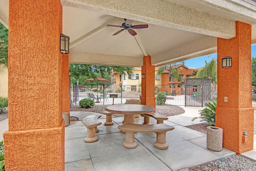 Covered patio here at The Retreat at Speedway Apartments with three stone picnic tables and benches on a concrete floor. Brick pillars surround the area, with a gate and apartment building in view.