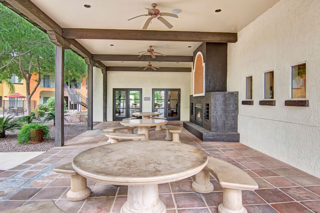 Covered outdoor patio here at The Retreat at Speedway Apartments with large stone tables and benches, ceiling fans, and a black fireplace. Adjacent to a lush garden and colorful building.