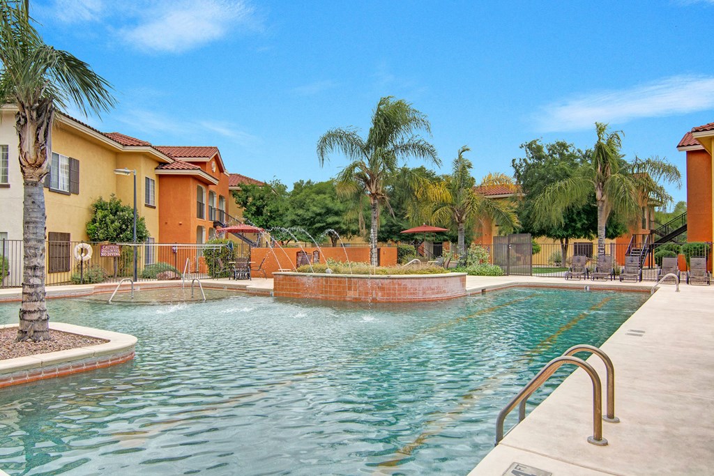 Sunny pool area here at The Retreat at Speedway Apartments with clear water and fountain jets surrounded by palm trees. Colorful buildings with red-tiled roofs frame the tranquil scene.