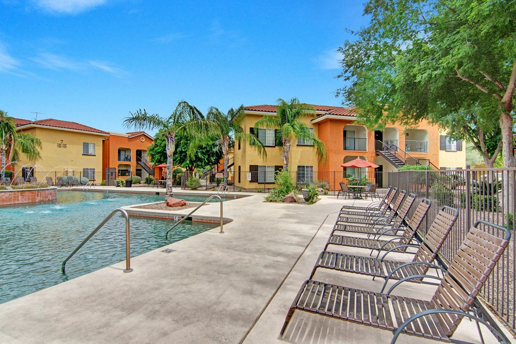 Poolside area here at The Retreat at Speedway Apartments with a calm pool and fountain, surrounded by yellow and orange apartment buildings. Palm trees and lounge chairs create a relaxed atmosphere.