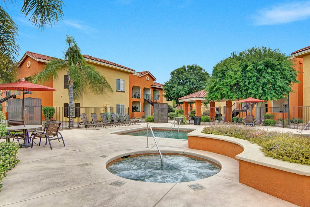 Sunny outdoor pool area here at The Retreat at Speedway Apartments with a jacuzzi, surrounded by lounge chairs, palm trees, and colorful buildings. Relaxed and inviting atmosphere.