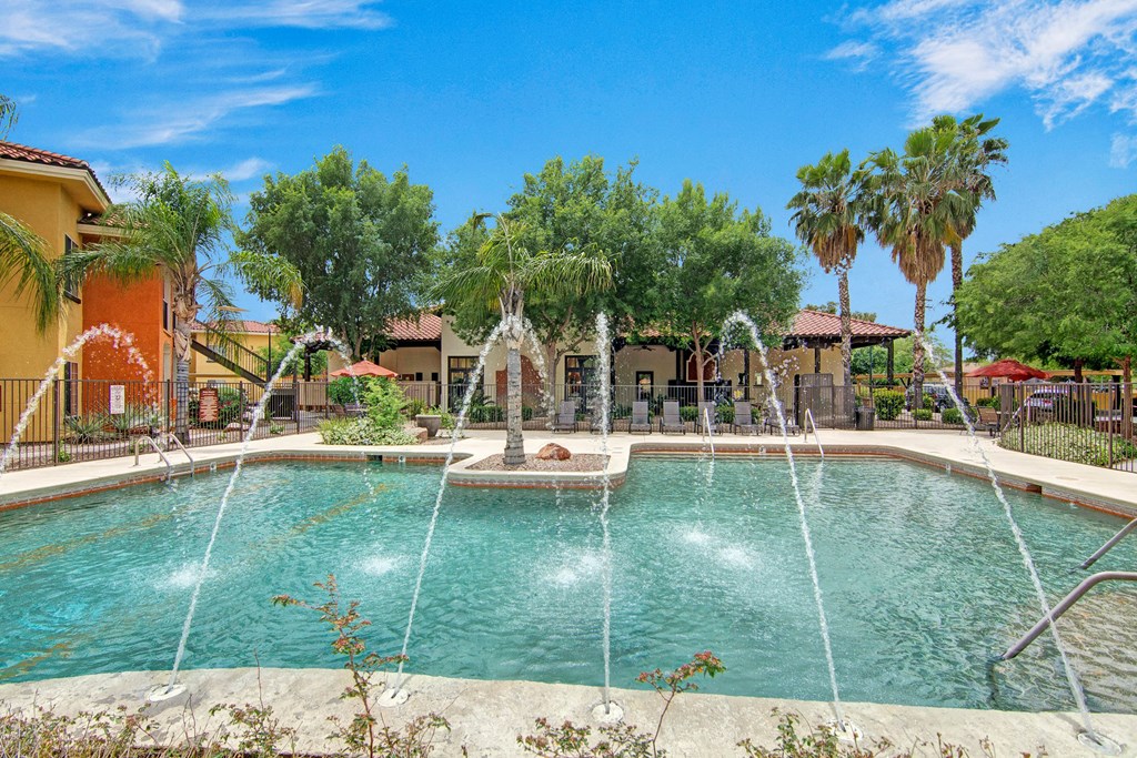 An outdoor pool with lounge chairs Outdoor pool here at The Retreat at Speedway Apartments with water fountains and clear blue sky, surrounded by tropical trees and colorful buildings, creating a relaxing, sunny atmosphere.