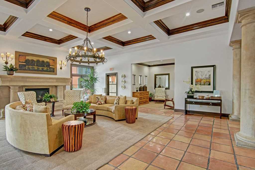 Elegant resident lounge here at The Retreat at Speedway Apartments with beige sofas, dark wood accents, and a large chandelier. A fireplace with a mantel, wall art, and plants add sophistication.