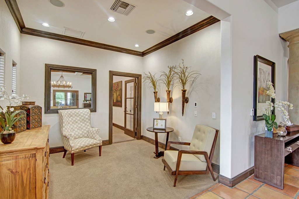Cozy, elegant waiting room here at The Retreat at Speedway Apartments with beige armchairs, a small round table with a lamp, wall-mounted floral decor, and framed art, creating a welcoming ambience.