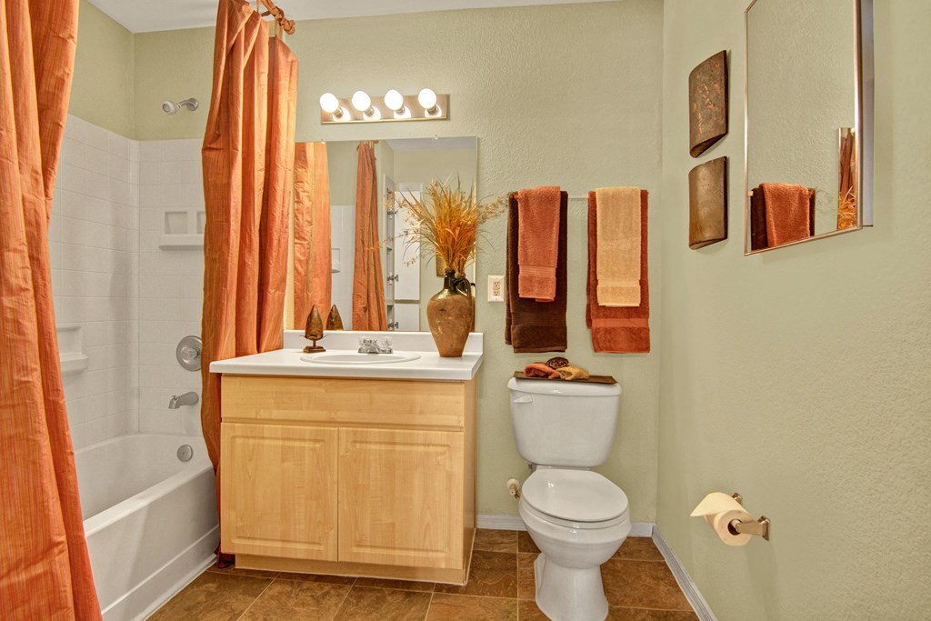 A cozy bathroom here at The Retreat at Speedway Apartments with a light wood vanity, white countertop, and modern faucet. Earth-toned towels hang near a toilet. An orange shower curtain complements the decor.