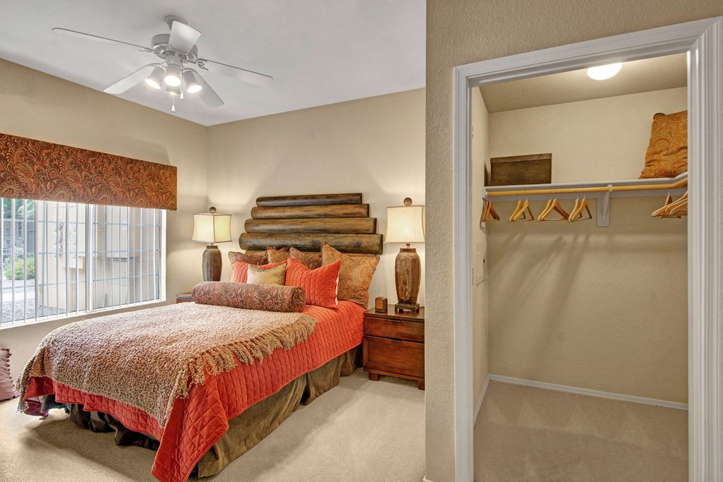 Retreat at Speedway Bedroom Cozy bedroom here at The Retreat at Speedway Apartments with wood headboard, red and beige bedding, and two lamps on nightstands. Open closet with hangers and a shelf on the right.