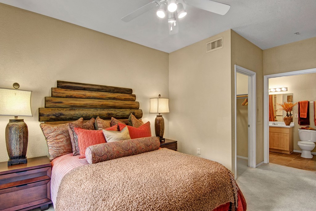 Cozy bedroom here at The Retreat at Speedway Apartments with a wood-paneled headboard, beige bedding, and decorative pillows. Warm lighting from bedside lamps enhances a calming ambiance. A door leads to a bathroom.