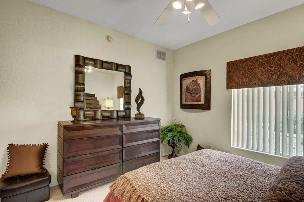 Cozy bedroom here at The Retreat at Speedway Apartments with a wooden dresser and mirror. A decorative sculpture and potted plant accent the space, with soft lighting creating a warm ambiance.
