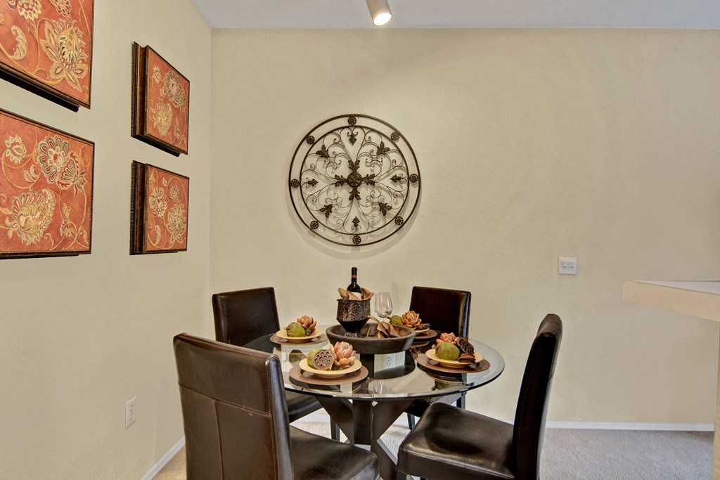 Dining area here at Retreat at Speedway Apartments with a glass round table set for four, featuring elegant centerpieces. Leather chairs surround the table. Decorative wall art and a metal wall piece add a sophisticated touch.