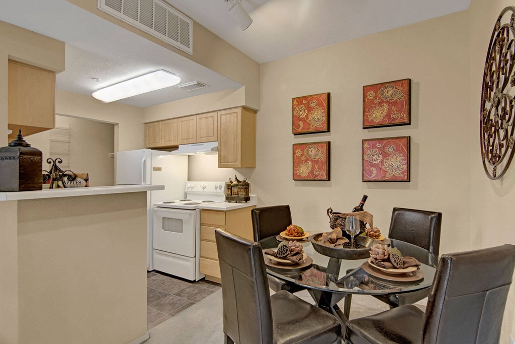 Modern kitchen-dining area here at Retreat at Speedway Apartments with beige walls, glass table, four black chairs, red floral wall art, white appliances, and elegant decor. Cozy ambiance.