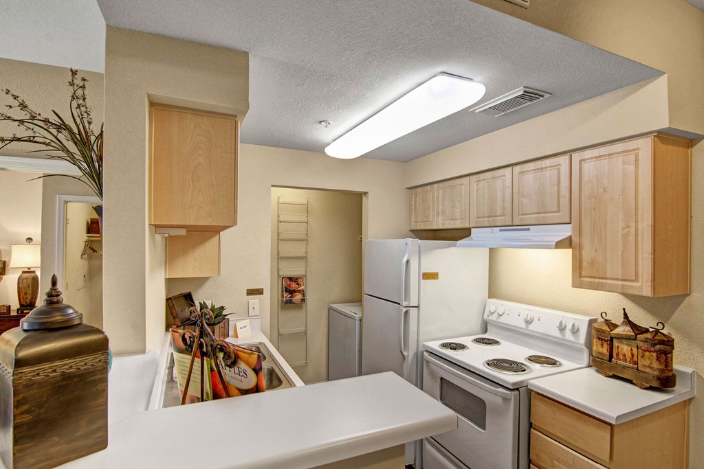 Compact kitchen here at Retreat at Speedway Apartments with light wood cabinets, white appliances, and a countertop with decorative items. Warm lighting creates a cozy atmosphere.