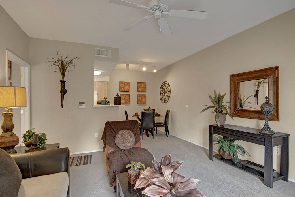 Modern living room here at The Retreat at Speedway Apartments with beige walls and carpet, featuring a ceiling fan, dark furniture, and decorative plants. A large mirror and artwork adorn the walls.
