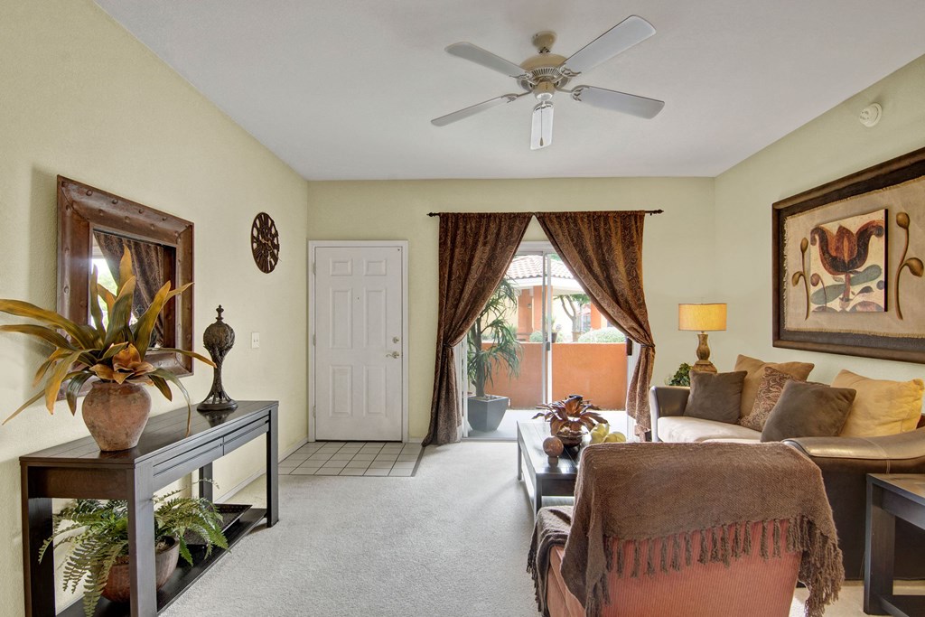A cozy living room here at The Retreat at Speedway Apartments with a beige sofa, floral artwork, and brown curtains. A ceiling fan hangs above; a console table with plants and lamps adds warmth.