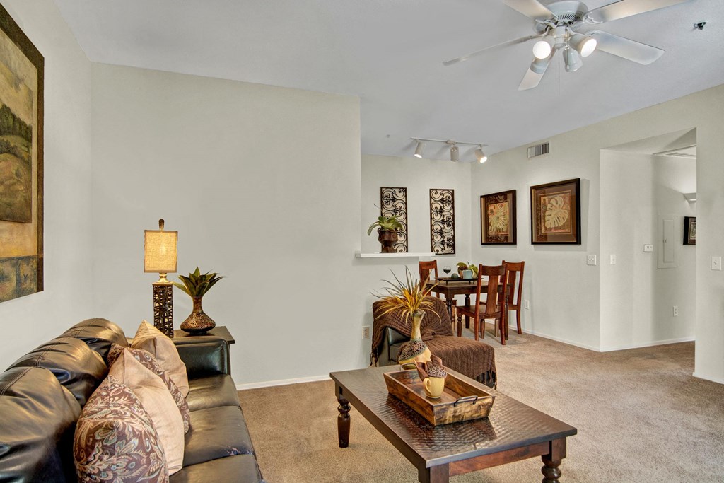 Spacious living room here at The Retreat at Speedway Apartments with a leather sofa, patterned pillows, and a table with plants. A cozy dining area is visible, decorated with nature-themed art. Neutral tones create a warm atmosphere.