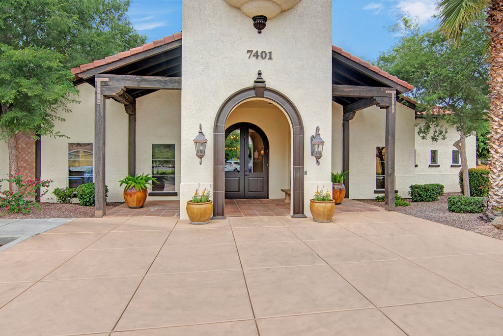 Entrance of our leasing office here at The Retreat at Speedway Apartments with an arched wooden door, flanked by lanterns. Potted plants line the entryway, surrounded by trees and lush greenery. Bright, inviting atmosphere.