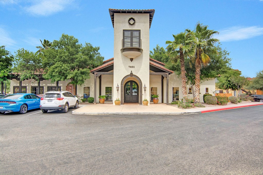Our Spanish-style leasing office here at The Retreat at Speedway Apartments with arched entrance and tower, surrounded by lush greenery and palm trees. Cars park nearby under a clear blue sky.