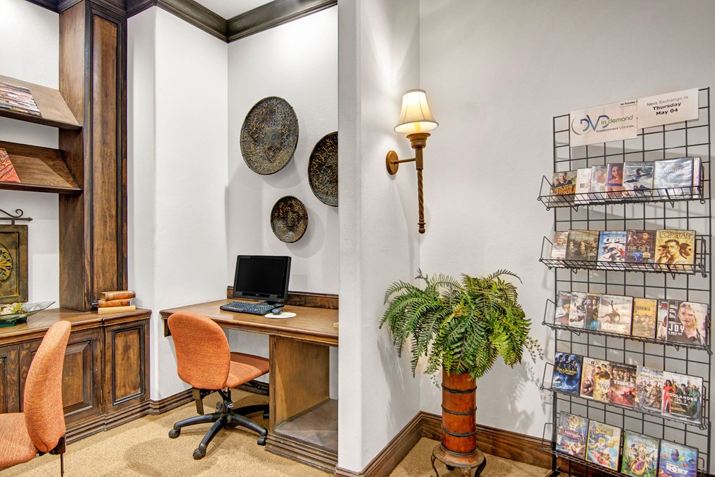 Cozy study area here at The Retreat at Speedway Apartments with a computer on a wooden desk, orange chairs, decorative metal plates on the wall, a fern in a pot, and a DVD rack.