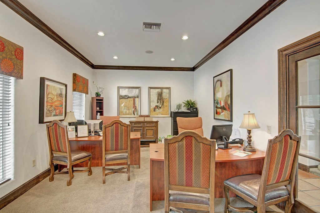 Warm office space here at The Retreat at Speedway Apartments with four wooden desks, high-backed chairs, and abstract art. Soft lighting from table lamps and natural light from windows. Cozy, professional ambiance.