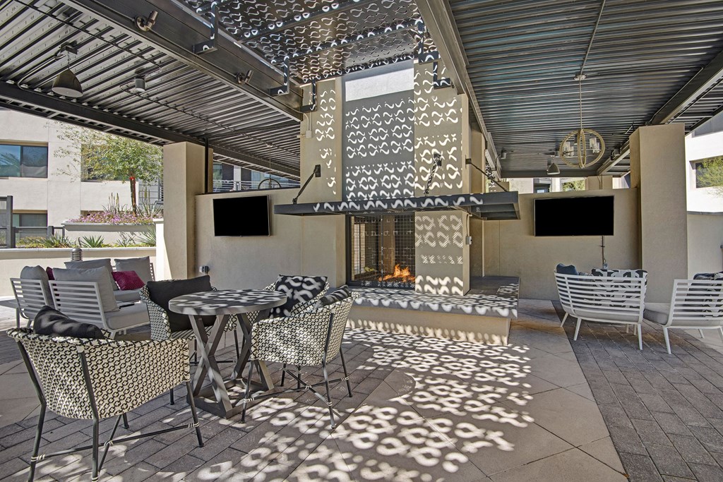 Modern outdoor patio here at Roosevelt Row Apartments with patterned shadows cast by metal pergola. Features cozy seating around a fireplace, large TVs, and a relaxed atmosphere.