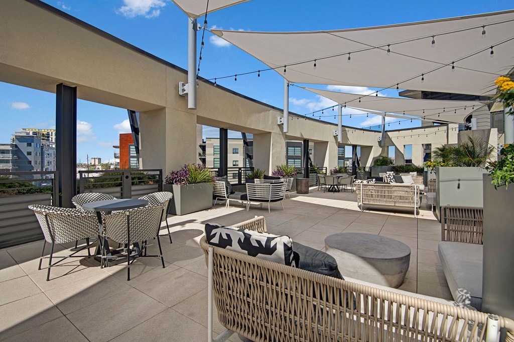 Rooftop terrace here at Roosevelt Row Apartments with modern seating, tables, and planters under white shade sails. String lights add ambiance. City skyline visible under a clear blue sky.