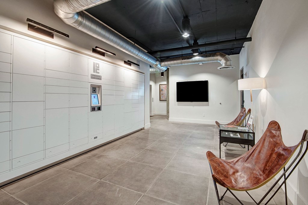 A modern lobby here at Roosevelt Row Apartments featuring white parcel lockers with a digital screen on the left. Two brown leather butterfly chairs and a lamp on the right, all under exposed ductwork.