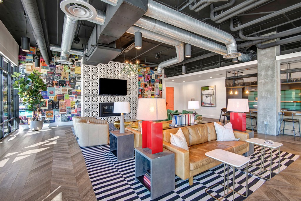 Modern lounge here at Roosevelt Row Apartments with eclectic decor featuring a tan leather sofa, colorful wall collage, striped rug, and exposed ducts. Bright, vibrant, and inviting atmosphere.