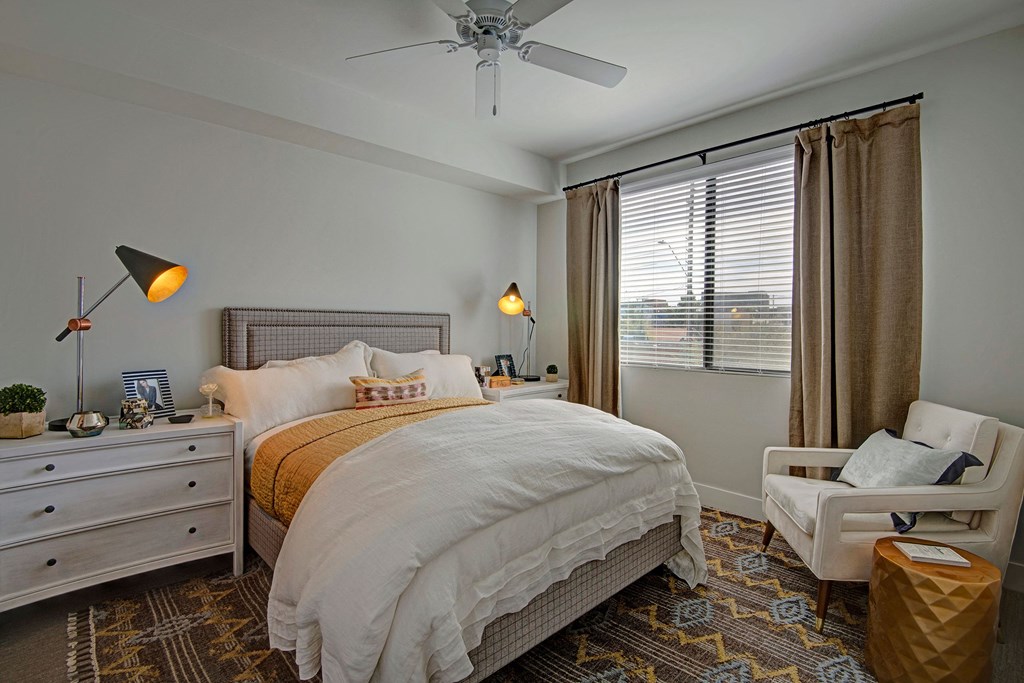 Roosevelt Row Bedroom Cozy bedroom here at Roosevelt Row Apartments with a large bed, white and mustard bedding, modern lamps, and a patterned rug. Natural light filters through blinds, creating a warm, inviting atmosphere.