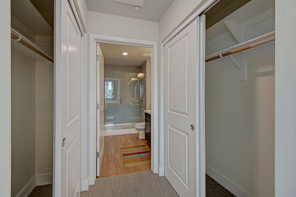 Hallway view here at Roosevelt Row Apartments of a white bathroom with clean lines and neutral tones. Dual closets flank the entry, leading to a glass shower and modern fixtures.