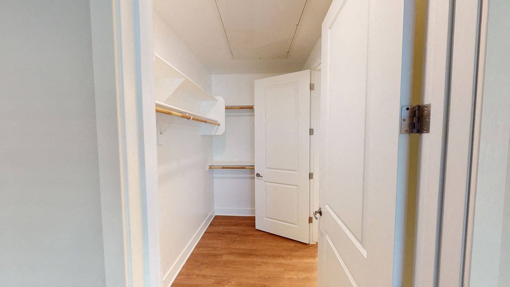 Narrow, empty walk-in closet here at Roosevelt Row Apartments with wooden floor, open white door, shelves, and hanging rods. The space feels clean, bright, and organized.