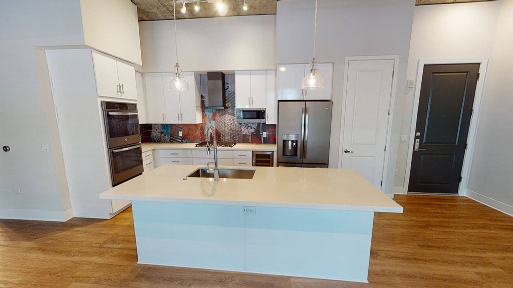 Modern kitchen here at Roosevelt Row Apartments with wooden floors, a large island with a sink, pendant lights, white cabinets, and colorful mosaic backsplash create a sleek look.