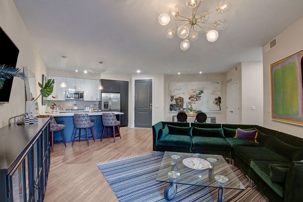 Modern living room here at Roosevelt Row Apartments with open kitchen. Green velvet sectional sofa, glass coffee table on striped rug, contemporary art, and a sleek chandelier overhead.