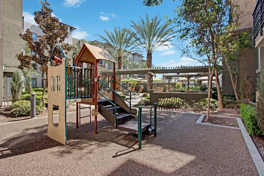 Playground here at Artisan at Main Street Metro Apartments with a small slide and steps in a sunlit courtyard, surrounded by lush greenery and palm trees, creating a peaceful and inviting atmosphere.