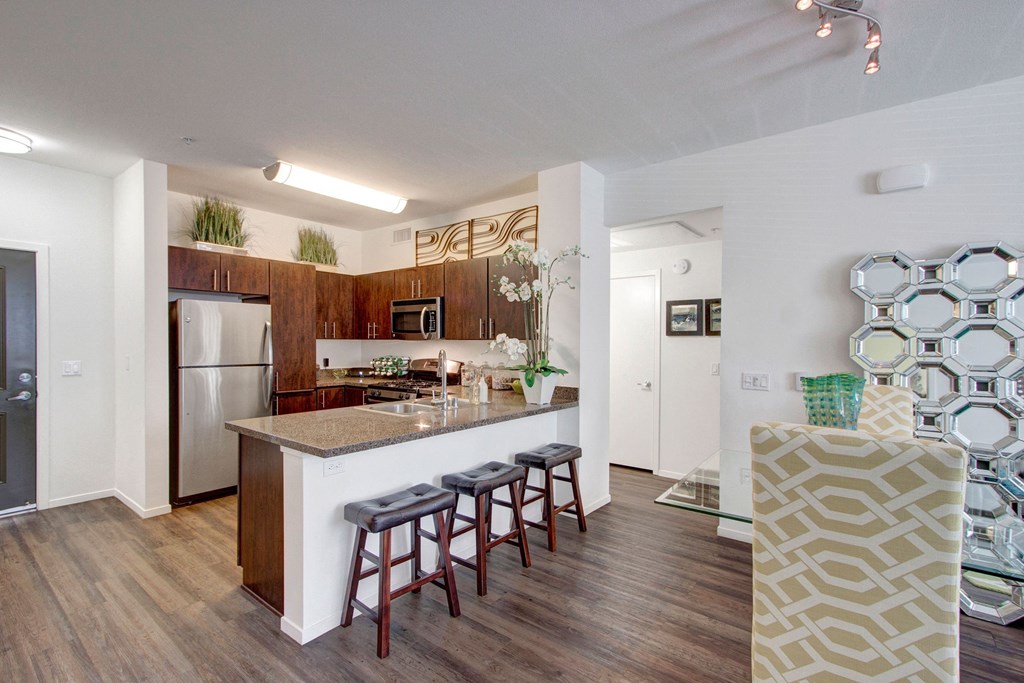 Modern kitchen here at Artisan at Main Street Metro Apartments with dark wood cabinets, stainless steel appliances, and a granite island featuring bar stools. Bright, airy, with decorative plants.
