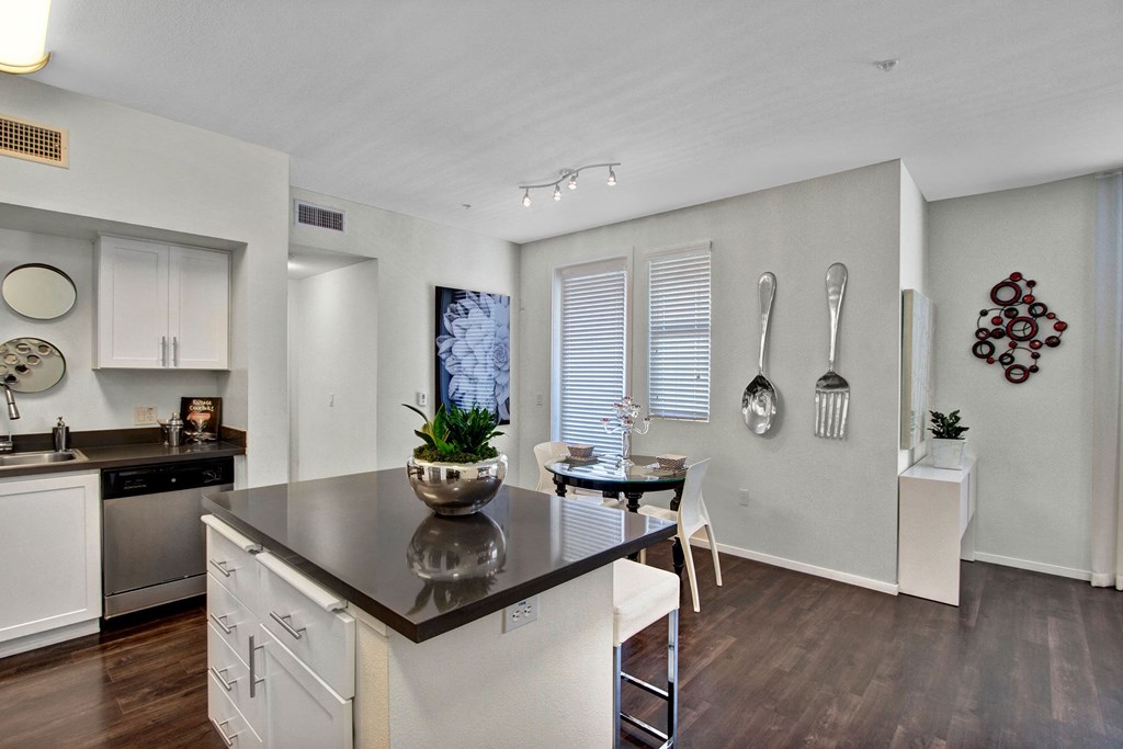 Modern kitchen and dining area here at Artisan at Main Street Metro Apartments with dark wood floors, white cabinets, and a gray countertop island. Wall decor includes oversized silver utensils.