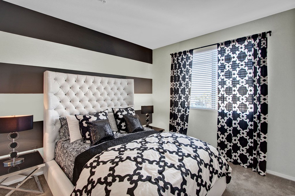 Sophisticated bedroom here at Artisan at Main Street Metro Apartments with gray and white striped walls, a tufted headboard, and matching patterned bedding and curtains. Two modern lamps add elegance.