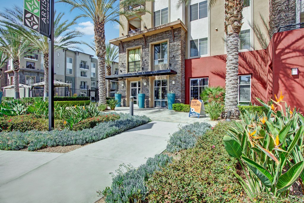 Exterior of our modern Artisan at Main Street Metro apartment complex with red accents, palm trees, and lush landscaping. A "Now Leasing" sign is displayed near the entrance.