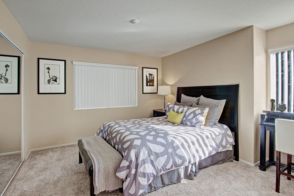 Castlepark Bedroom Cozy bedroom here at Castlepark Apartment Homes with a modern design. Features a bed with patterned gray and white bedding, a bedside lamp, abstract framed art, and a small desk by the window.
