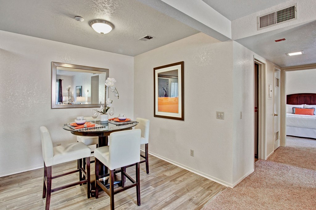 Castlepark Dining Room Cozy dining area here at Castlepark Apartment Homes with a round glass table, white chairs, and an orchid centerpiece. Mirror on one wall; adjacent to a hallway leading to a bedroom. Warm, inviting tone.