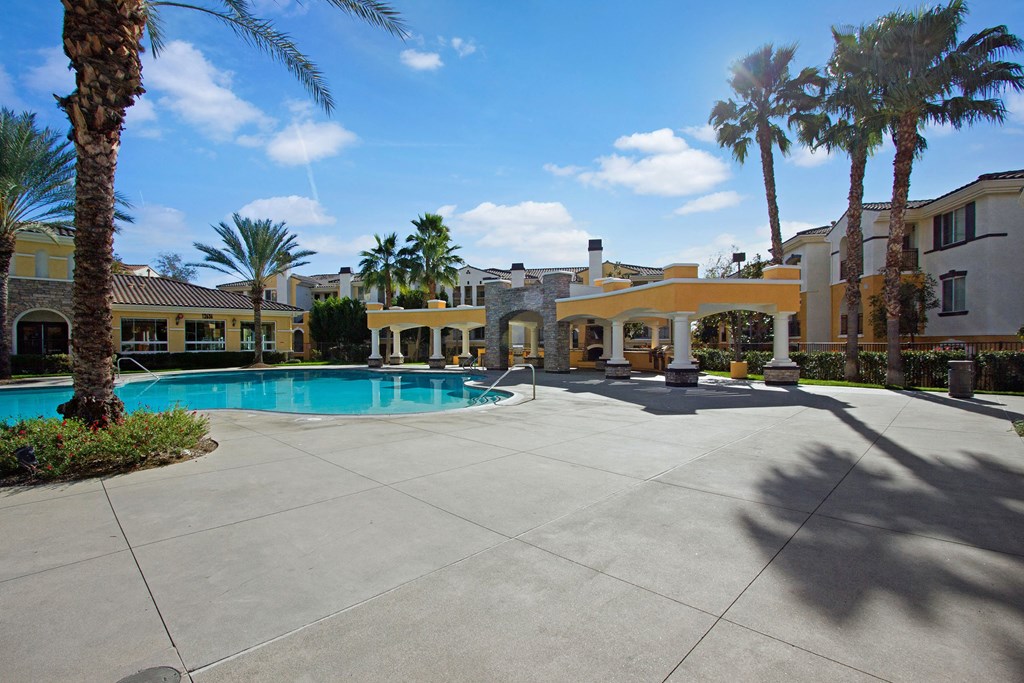 Sunny courtyard here at Fresco Apartment Homes with a clear blue pool surrounded by palm trees and patio seating, adjacent to an elegant building with arched architecture.