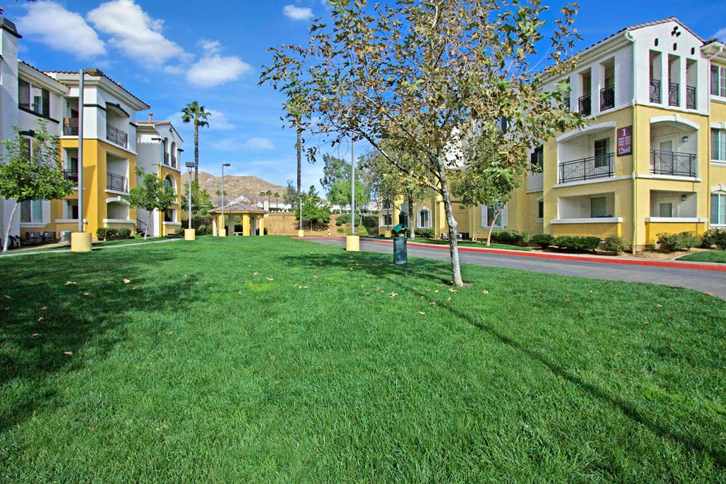 Spacious courtyard here at Fresco Apartment Homes with vibrant green grass flanked by yellow and white apartment buildings under a bright blue sky. Trees and mountains in the background.