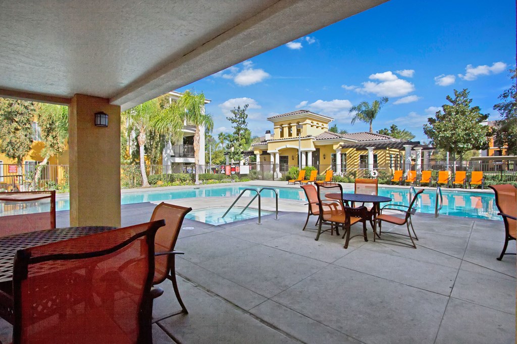 Covered patio here at Fresco Apartment Homes overlooking a sunny pool with lounge chairs and a clubhouse surrounded by palm trees. The scene is peaceful and inviting.