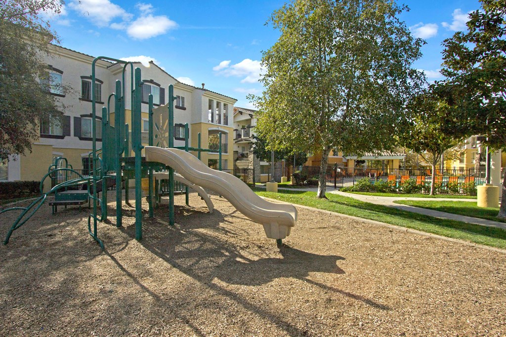 Playground with a slide and climbing structure on wood chips here at Fresco Apartment Homes, adjacent to a residential building. Trees and pathways are in a sunny, peaceful setting.