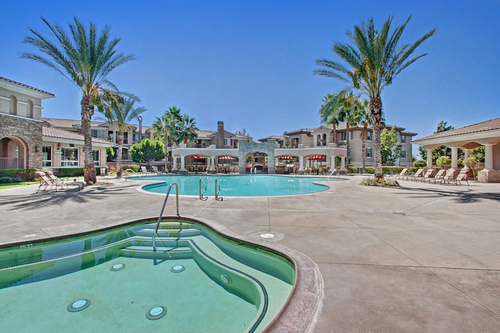 Luxurious outdoor pool area here at Fresco Apartment Homes with palm trees in sunny weather. Features a hot tub in the foreground, lounge chairs, and elegant buildings in the background.