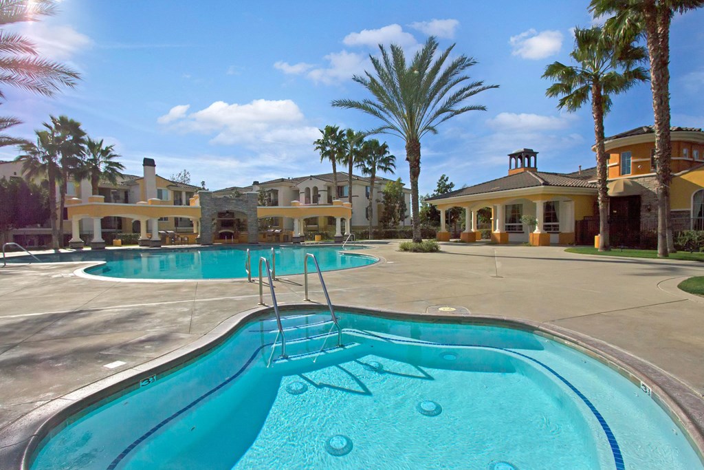 Sunny poolside view here at Fresco Apartment Homes featuring a tranquil jacuzzi in the foreground and large pool surrounded by palm trees and resort-style buildings in the background.