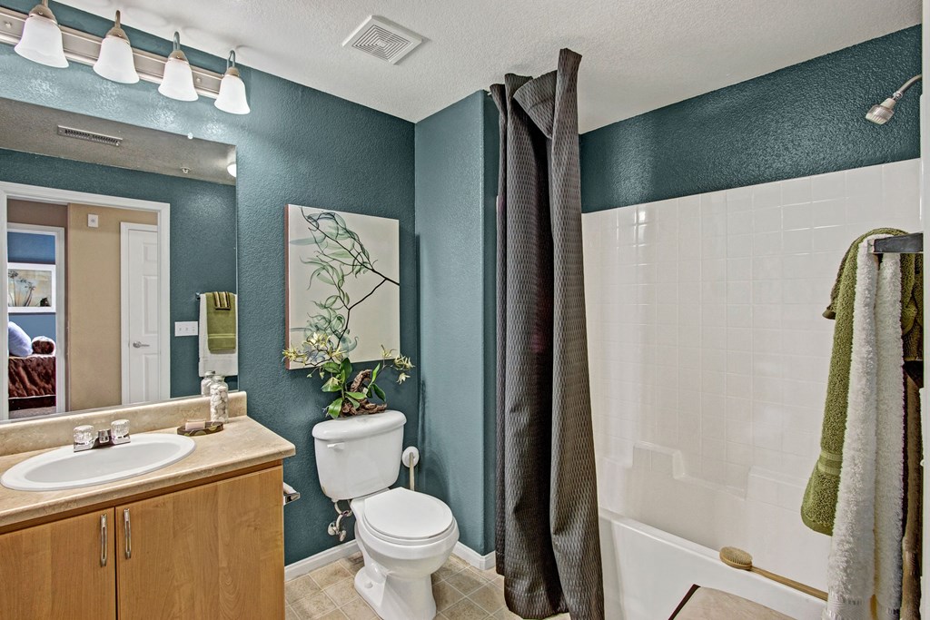 A bathroom here at Fresco Apartment Homes with teal walls features a wood cabinet sink, large mirror, and modern lighting. A toilet and a shower with dark curtains are visible.
