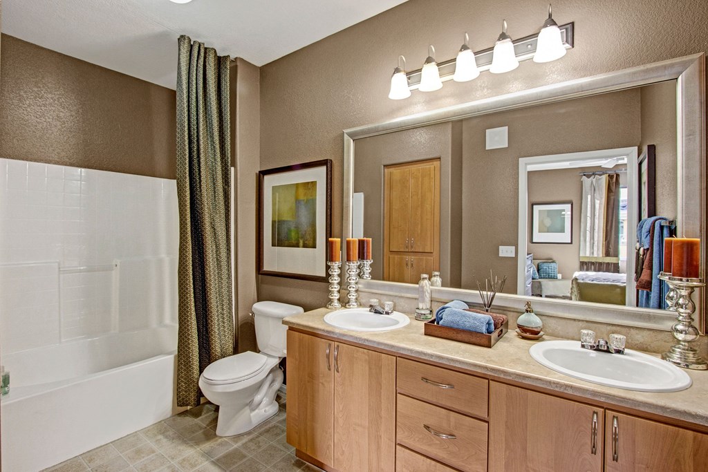 Elegant bathroom here at Fresco Apartment Homes with dual sinks, large mirror, and soft lighting. Features a bathtub with a patterned curtain, modern decor, and earthy colors. Relaxing ambiance.