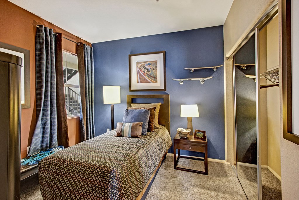 A cozy bedroom here at Fresco Apartment Homes with a single bed adorned with patterned bedding and several cushions. The room features blue and brown walls, two illuminated lamps, a nightstand, and decorative skateboard shelves. A framed painting hangs above the bed, adding a touch of elegance. The atmosphere is warm and inviting.