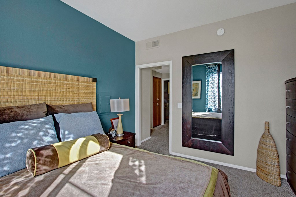 A cozy bedroom here at Fresco Apartment Homes with beige and teal walls, featuring a wicker headboard and plush bed with blue and brown pillows. Sunlight casts leaf shadows across the bedspread. A sleek, dark-framed mirror reflects a patterned curtain. A wicker vase and a small nightstand with a lamp add warmth.
