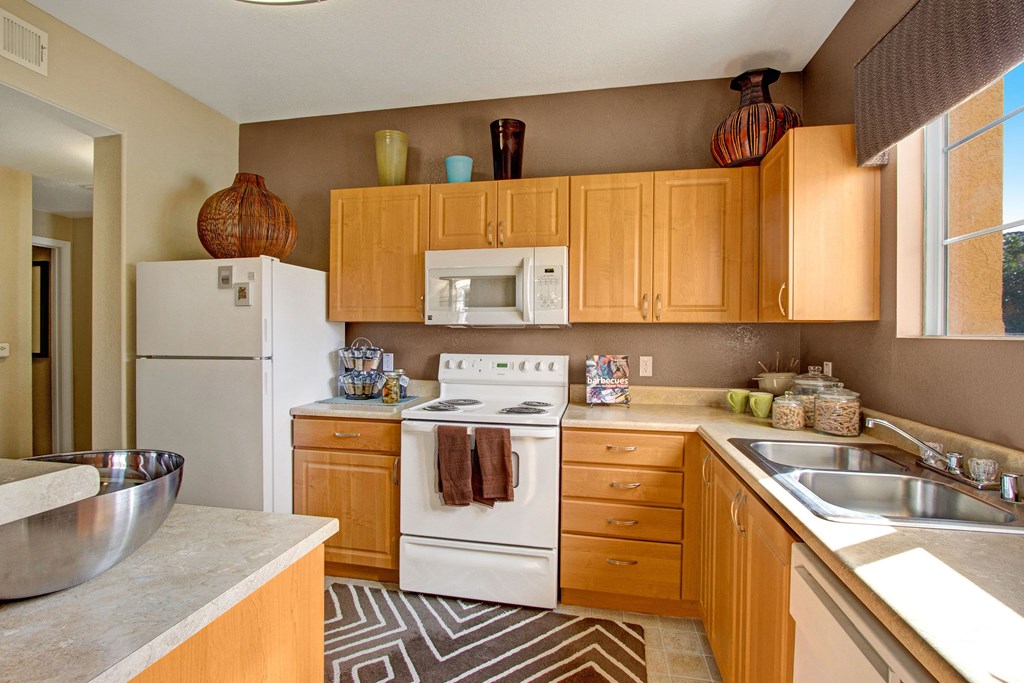 Modern kitchen here at Fresco Apartment Homes with wooden cabinets, white appliances, and a double sink. Decorative vases on top, brown and white patterned rug, and sunlit window.